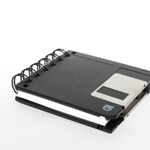 May include: A black spiral notebook with a floppy disk attached to the cover. The floppy disk is silver and has a black label with the letter "D" on it.