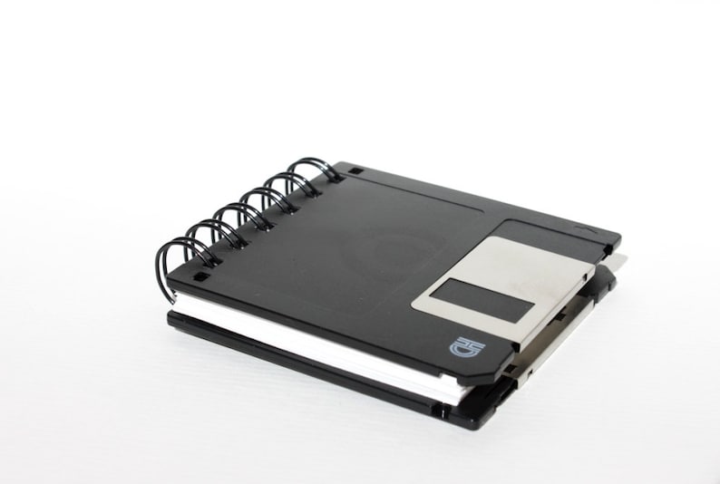 May include: A black spiral notebook with a floppy disk attached to the cover. The floppy disk is silver and has a black label with the letter "D" on it.