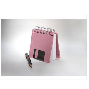 May include: A pink floppy disk shaped notebook with a silver spiral binding. The notebook is closed and sitting on a white surface with a black pen next to it.