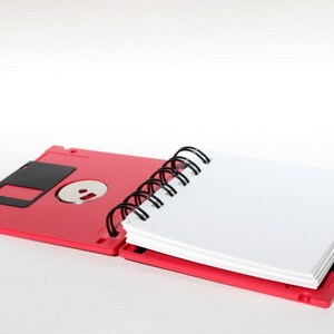 May include: A red notebook with a spiral binding. The notebook is made to look like a floppy disk. The notebook is open and the pages are blank.