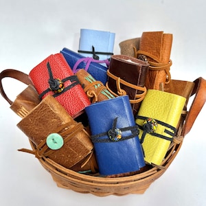 May include: A basket filled with small leather-bound journals in various colors, including blue, brown, red, yellow, and green. Each journal is tied with a leather strap and has a decorative button.