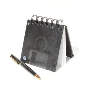 May include: A black spiral bound notebook with a floppy disk design. The notebook is open and a black pen is lying next to it.