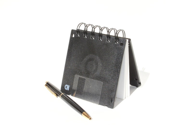 May include: A black spiral bound notebook with a floppy disk design. The notebook is open and a black pen is lying next to it.