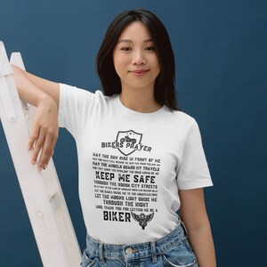 May include: White t-shirt with a black print of a biker prayer. The text reads "Bikers Prayer" and includes a motorcycle graphic. The text continues with "May the sun rise in front of me, May the rain fall behind me, May the angels guard my travels, Keep me safe through the rough city streets, Let the hogs light guide me, Let the roads lead me the right way, Thank you for letting me be a biker."