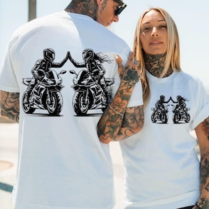 May include: Two white t-shirts with a black graphic of two people riding motorcycles and high-fiving each other.