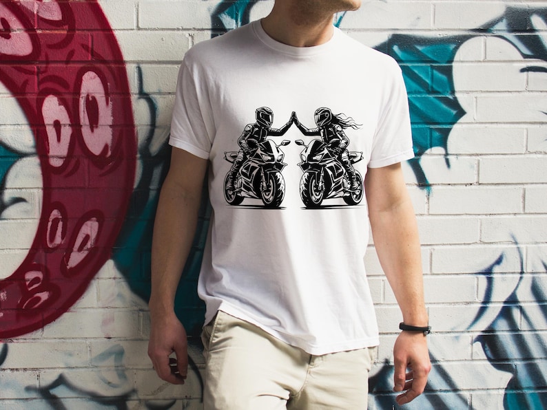 May include: White t-shirt with a black and white graphic of two people riding motorcycles. The graphic shows the riders giving each other a high five.