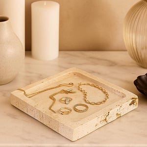 May include: A square, beige stone jewelry tray with gold necklaces, rings, and a bracelet. The tray sits on a marble surface, with a vase, candles, and decorative objects in the background. The jewelry is displayed on the tray.