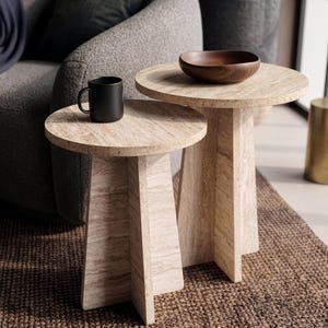 May include: Two round side tables made of beige travertine stone with a natural veined pattern. The tables feature a pedestal base with four intersecting panels. A black ceramic mug sits on the smaller table, and a dark wooden bowl sits on the taller table.