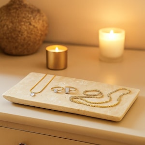 May include: A gold-toned jewelry set displayed on a rectangular, light-colored stone tray. The set includes a necklace with a round pendant, two rings, and a delicate chain bracelet. Two lit candles and a decorative vase are in the background.
