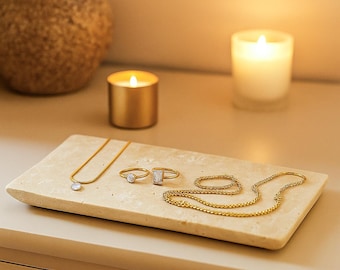May include: A gold-toned jewelry set displayed on a rectangular, light-colored stone tray. The set includes a necklace with a round pendant, two rings, and a delicate chain bracelet. Two lit candles and a decorative vase are in the background.