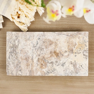 Könnte beinhalten: Eine rechteckige Steinplatte mit strukturierter Oberfläche. Sie zeigt eine Mischung aus Beige-, Grau- und Brauntönen mit dunkleren Adern. Im Hintergrund befinden sich ein cremefarbenes Handtuch mit Fransen und weiße Orchideenblüten. Die Platte ist ca. 30 cm lang.