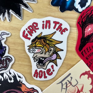 May include: A white sticker with a cartoon character with yellow hair, goggles, and a long tongue. The character is saying "Fire in the hole!" in red letters.