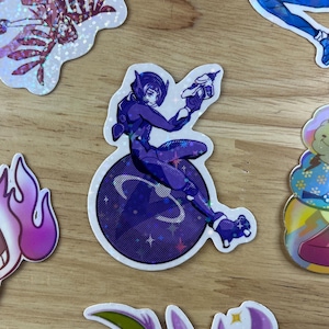 Peut inclure: Un autocollant holographique représentant un personnage vêtu de violet tenant une arme à feu, debout sur une planète violette et bleue avec des étoiles. L'autocollant est sur une surface en bois.