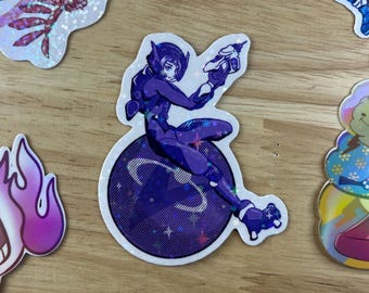May include: A holographic sticker featuring a purple-clad character holding a gun, standing on a purple and blue planet with stars. The sticker is on a wooden surface.