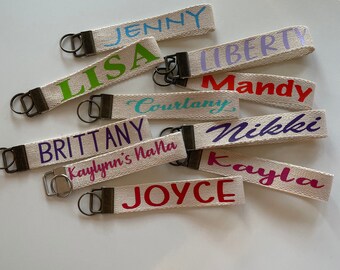 May include: A collection of ten personalized keychains with various names printed on them in different colors. The keychains are made of beige fabric with a metal ring.