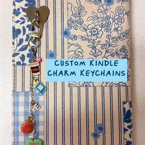 May include: A Kindle cover with a blue and white floral and striped pattern, featuring a heart-shaped clasp and a charm keychain. The keychain includes charms of a coffee cup, a book, a seashell, and a green heart with the text "Sweet Heart". The text "CUSTOM KINDLE CHARM KEYCHAINS" is visible.