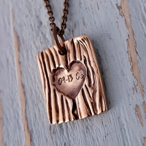 Op de afbeelding: Een koperen ketting met een vierkante hanger met een hart met de datum "01-13-03" erin gegraveerd. De hanger heeft een getextureerd, hout-achtig oppervlak.