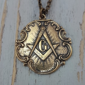 May include: A gold-toned pendant necklace featuring a detailed ornate design with a compass and square symbol, and the letter 'G' in the center.