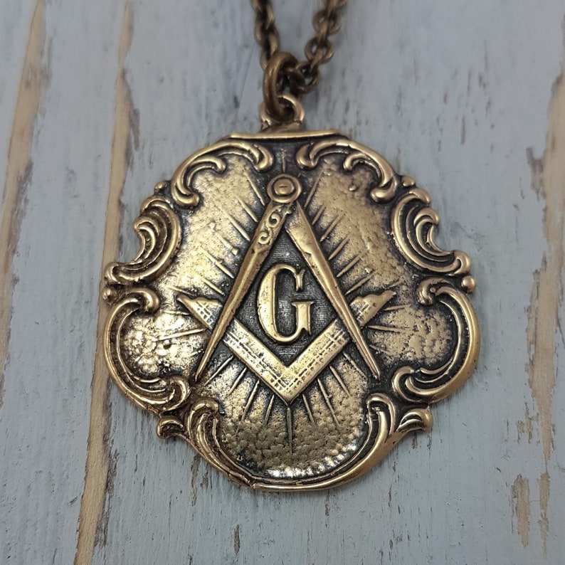 May include: A gold-toned pendant necklace featuring a detailed ornate design with a compass and square symbol, and the letter 'G' in the center.