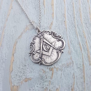 May include: Silver necklace with a pendant featuring a Masonic symbol, a compass and square with a 'G' in the centre, surrounded by a decorative frame.