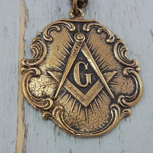 May include: A gold-toned pendant featuring a Masonic compass and square with a 'G' in the center. The pendant has a decorative, ornate border.
