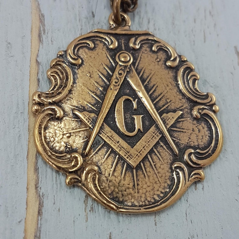 May include: A gold-toned pendant featuring a Masonic compass and square with a 'G' in the center. The pendant has a decorative, ornate border.