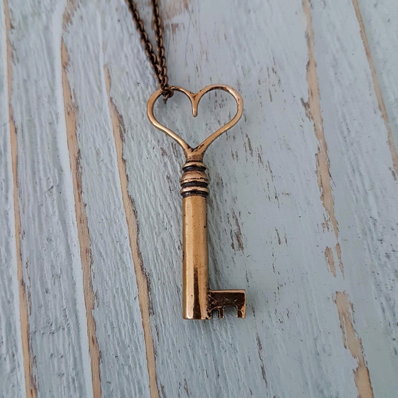 Op de afbeelding: Een gouden sleutelketting met een hartvormig sleutelgat. De sleutel hangt aan een ketting en aan een houten oppervlak.