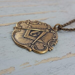 May include: A gold-toned pendant necklace featuring a detailed ornate design with a compass and square symbol, and the letter "G" in the center.