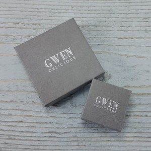 Op de afbeelding: Twee grijze dozen met de tekst "GWEN DELICIOUS" in wit gedrukt op de bovenkant van elke doos.