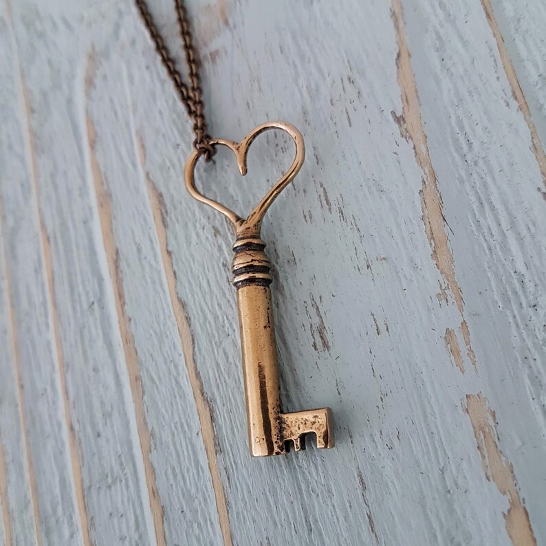 Op de afbeelding: Een gouden ketting met een sleutel in de vorm van een hart. De sleutel hangt aan een ketting en aan een houten oppervlak.