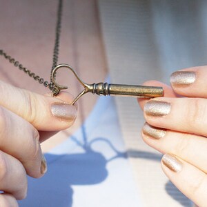 Op de afbeelding: Een gouden sleutel hanger ketting met een hartvormige lus. De sleutel zit aan een ketting en de persoon houdt de hanger in zijn handen. De vingernagels van de persoon zijn gelakt met een glinsterende gouden nagellak.