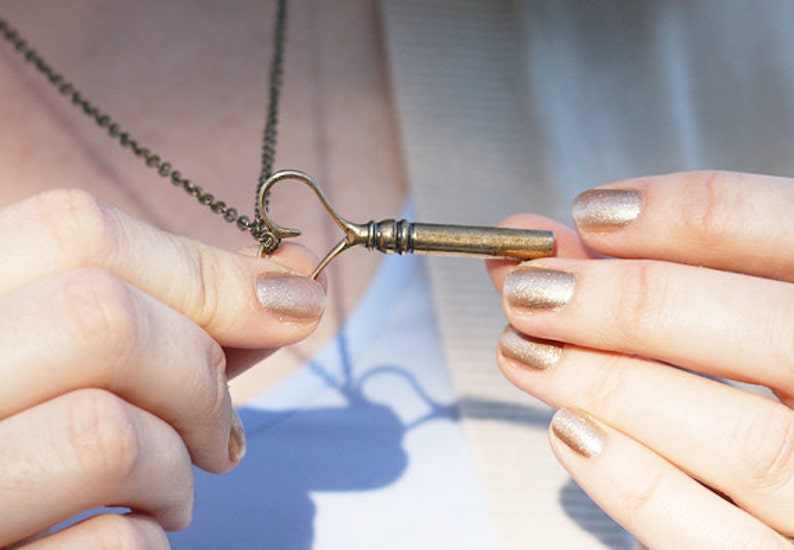 Op de afbeelding: Een gouden sleutel hanger ketting met een hartvormige lus. De sleutel zit aan een ketting en de persoon houdt de hanger in zijn handen. De vingernagels van de persoon zijn gelakt met een glinsterende gouden nagellak.
