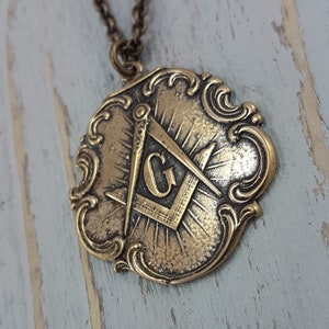 May include: A gold-toned pendant necklace featuring a detailed, ornate design with a Masonic symbol, including a compass and square, and the letter 'G' in the centre.