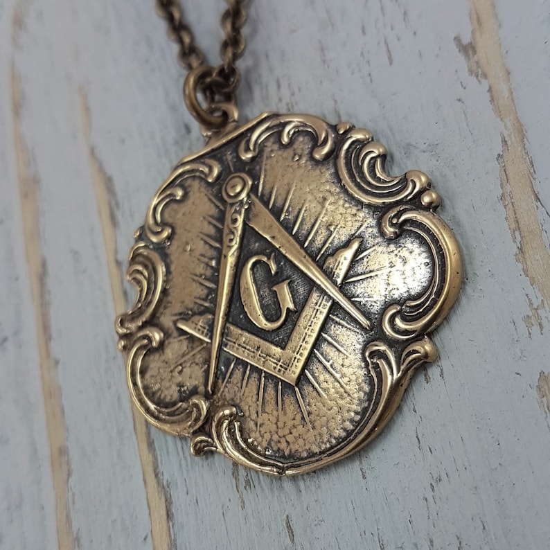 May include: A gold-toned pendant necklace featuring a detailed, ornate design with a Masonic symbol, including a compass and square, and the letter 'G' in the center.