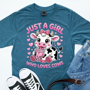 May include: Teal t-shirt with a cartoon cow drinking a pink drink, text reads "JUST A GIRL WHO LOVES COWS". The cow is black and white with a pink bow. The shirt is paired with jeans and black and white trainers.