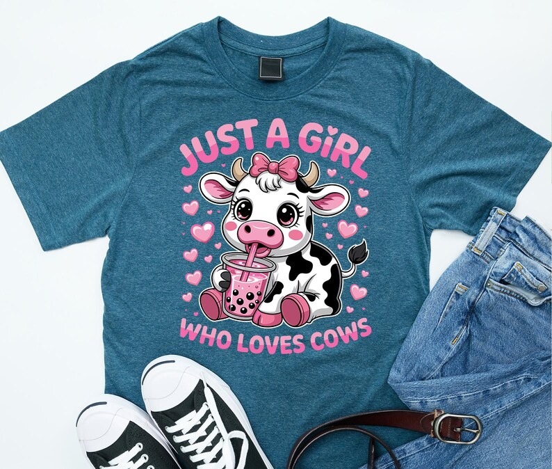 May include: Teal t-shirt with a cartoon cow drinking a pink drink, text reads "JUST A GIRL WHO LOVES COWS". The cow is black and white with a pink bow. The shirt is paired with jeans and black and white trainers.
