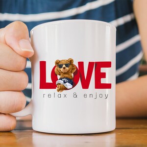 May include: White ceramic mug with the red word "LOVE" and a cartoon bear wearing sunglasses and shorts. The text "relax & enjoy" is printed below the bear. The mug is held by a person.