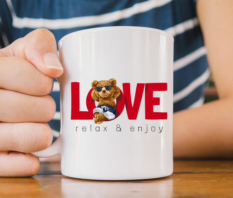 May include: White ceramic mug with the red word "LOVE" and a cartoon bear wearing sunglasses and shorts. The text "relax & enjoy" is printed below the bear. The mug is held by a person.