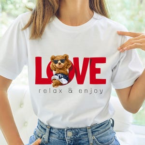 May include: White t-shirt featuring a red "LOVE" graphic and a cartoon bear wearing sunglasses and swim trunks. The text "relax & enjoy" is below the graphic. The shirt is made of a soft material and is suitable for casual wear.