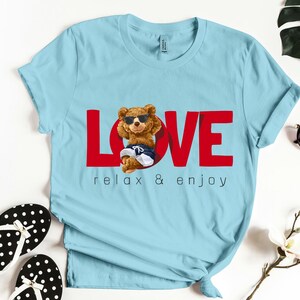 May include: Pale blue t-shirt with the word "LOVE" in red, featuring a cartoon bear wearing sunglasses and swim shorts. The text "relax & enjoy" is below the word "LOVE".