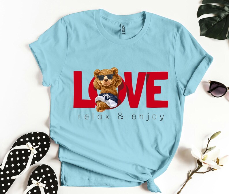 May include: Pale blue t-shirt with the word "LOVE" in red, featuring a cartoon bear wearing sunglasses and swim shorts. The text "relax & enjoy" is below the word "LOVE".