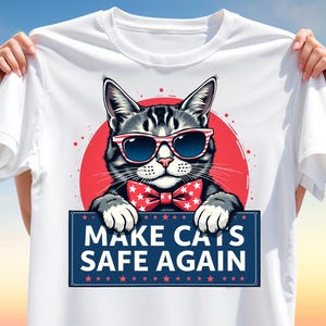 May include: White t-shirt featuring a cat wearing sunglasses and a bow tie, set against a red circle. The cat holds a sign that reads "MAKE CATS SAFE AGAIN" in blue and white. The design has a patriotic theme.