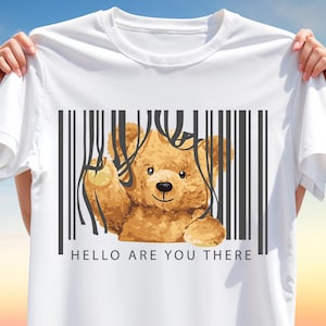 Puede incluir: Camiseta blanca con un diseño de oso de peluche de dibujos animados. El oso marrón está detrás de un gráfico de código de barras negro. El texto "HELLO ARE YOU THERE" está impreso debajo. La camiseta es sostenida por una persona.