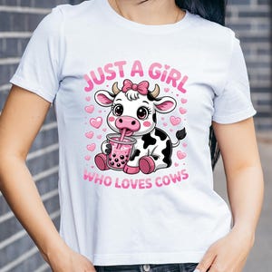 May include: White t-shirt with a cartoon cow design. The cow has a pink bow and is drinking from a pink drink. Pink text reads "JUST A GIRL WHO LOVES COWS". Pink hearts are also present.