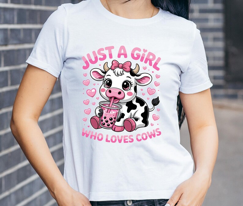 May include: White t-shirt with a cartoon cow design. The cow has a pink bow and is drinking from a pink drink. Pink text reads "JUST A GIRL WHO LOVES COWS". Pink hearts are also present.