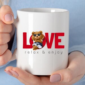 May include: White ceramic mug featuring the word "LOVE" in red block letters. A cartoon bear wearing sunglasses and swim shorts is in the centre. The phrase "relax & enjoy" is printed below the word "LOVE".