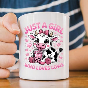 May include: White ceramic mug with a cartoon cow design. The cow, with a pink bow, is drinking a pink drink. The mug has the text "JUST A GIRL WHO LOVES COWS" in pink. The cow has black and white spots and is surrounded by pink hearts.