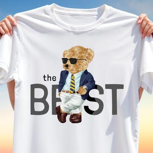 May include: White t-shirt featuring a cartoon bear wearing sunglasses, a navy blue blazer, and a yellow striped tie. The text "the BEST" is printed below the bear. The shirt is held up by hands.
