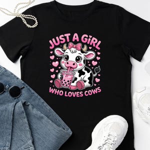 May include: Black t-shirt with a cartoon cow design, drinking a pink drink, and the text "JUST A GIRL WHO LOVES COWS" in pink. The design includes pink hearts. The shirt is shown with blue jeans, sunglasses, and white trainers.