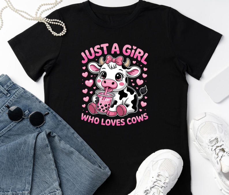 May include: Black t-shirt with a cartoon cow design, drinking a pink drink, and the text "JUST A GIRL WHO LOVES COWS" in pink. The design includes pink hearts. The shirt is shown with blue jeans, sunglasses, and white trainers.